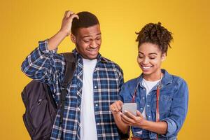 African Girlfriend Showing Travel App On Smartphone To Boyfriend Preparing For Vacation Standing Over Yellow Studio Background. Mobile Application And Service For Tourists Concept photo