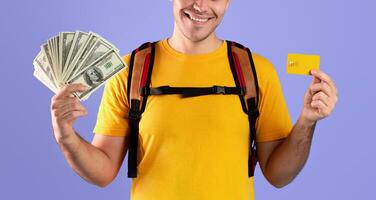 Planning travel budget concept. Portrait of funky young man with backpack, credit card and money smiling and looking at camera over blue studio background photo