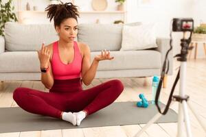Fitness Blogging Concept. African American Blogger Girl Making Workout Explaining How To Exercise And Stay Fit At Home, Sitting On Gymnastics Mat In Front Of Camera. Selective Focus photo