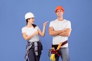Cheerful Couple In Hard Hats And Casual Clothes Having Fun During Repair, Excited Smiling Woman Pointing At Man With Tool Belt While They Standing Together Over Blue Studio Background, Copy Space photo