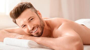 Happy middle aged man attending spa salon on weekend, lying on his belly on massage table, closeup. Handsome bearded man smiling at camera while waiting for relaxing back massage photo