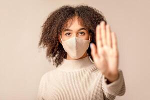 Woman suffer from sick, wearing mask and comply with social distance. Focus on face of african american lady in protective mask, fenced off with hand, isolated on light background, studio shot photo