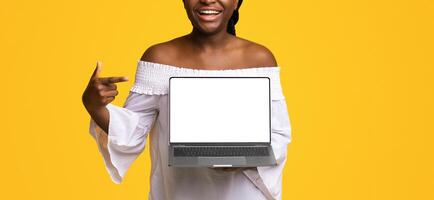 Check This Website. Cheerful Black Lady Pointing At Laptop Computer With Black Blank Screen, Standing On Yellow Background, Showing Free Place For Advertisement Or Design, Mockup With Copy Space photo