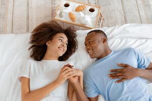 Happy morning, romantic breakfast and tenderness couple at home. Smiling young african american wife and husband lie on soft white bed in bedroom interior with tray with food and coffee, top view photo