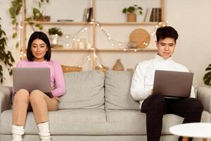 Korean Couple Using Laptop Computers Sitting On Couch At Home On Weekend Evening. Online Relationship Concept, Internet Websites And Family Web Life. Free Space For Text photo