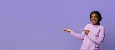 Joyful Black Millennial Woman Pointing Finger At Open Palm, Posing Over Purple Studio Background, Holding Invisible Object, Demonstrating And Recommending Something, , Panorama With Copy Space photo