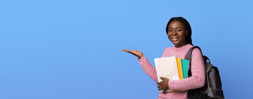 Smiling african american female student pointing aside at copy space on purple background, holding workbooks and backpack, demonstrating invisible object on her palm, recommending something, panorama photo