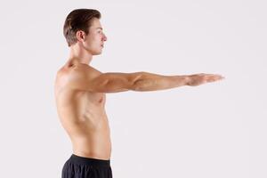 Side view of young strong guy with naked torso and prominent muscles doing aerobic warm up on light studio background. Handsome millennial sportsman making squats exercises photo