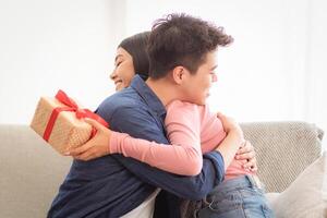 Romantic Gift. Korean Couple Hugging Holding Wrapped Present Box, Celebrating Relationship Anniversary Or Family Holiday Sitting On Couch At Home. Birthday Celebration Concept. Panorama photo