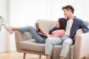 Asian Couple Using Laptop Watching Movie Online Relaxing Together Sitting On Couch At Home On Weekend. Modern Lifestyle, Computer Users Browsing Internet Concept. Free Space For Text photo