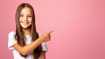 Wow, cool. Smiling little child points with finger behind back, to empty space and presents product, isolated on pink background photo