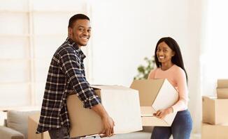 Black millennial couple carrying carton boxes in their new apartment on moving day. African American homeowners with belongings smiling and looking at camera, panorama photo