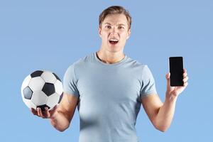 Young man with soccer ball disappointed over defeat of his favorite football team, watching match online on cellphone, violet studio background. Mockup for mobile app design photo