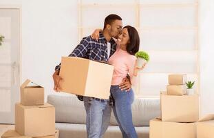 Affectionate black guy kissing and hugging his wife, holding carton box and house plant on moving day, panorama. Loving man and his girlfriend unpacking belongings in their new apartment photo