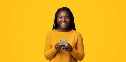Cool App. Cheerful African Female Holding Smartphone And Smiling At Camera, Browsing Internet Or Texting Wiith Friends, Checking New Application, Wearing Sweater, Posing Over Yellow Studio Background photo