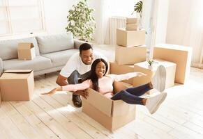 Beautiful black lady and her young husband playing together with cardboard box on moving day. Positive African American family entertaining themselves in their new house during relocation photo