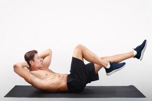 Strong young guy with bare torso doing abs exercises on yoga mat against light studio background, side view. Full length portrait of handsome athletic millennial man working out his core muscles photo
