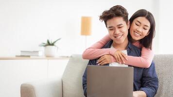 Happy Korean Couple Using Laptop Computer Relaxing Watching Movie Online Sitting On Sofa At Home. Asian Family Enjoying Weekend Together Browsing Internet Indoor. Free Space For Text photo