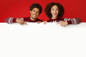 Happy New Year and merry Christmas. Laughing young african american couple in love in identical sweaters over white blank billboard and point fingers on it, isolated on red background, studio shot photo