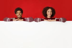 New Year and Christmas discounts and sales. Smiling young african american husband and wife in same clothes lean their hands on empty white billboard for words, isolated on red background, studio shot photo