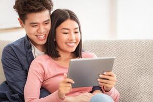 Happy Japanese Couple Using Digital Tablet Watching Movie Online Sitting At Home. Girlfriend And Boyfriend Relaxing Computer Using Computer, Spending Weekend Together. Gadgets And Modern Lifestyle. photo