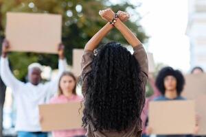 Unrecognizable long-haired brunette woman leading group of protestors holding blank placards, back view of lady standing in front of multiracial demonstrators, raising fists up and crossing hands photo