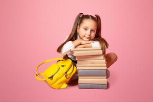 New knowledge for little schoolgirl. Child with backpack sits near stack of books isolated on pink background, empty space photo