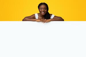 Place For Your Ad. Joyful Black Woman Leaning On Blank White Advertisement Board With Free Copy Space For Text Or Design, Standing Behind Blank Vertical Poster Over Yellow Studio Background photo