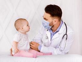 Doctor Pediatrician In Face Mask Examining Baby Patient Having Medical Checkup In Hospital Indoors. Sick Little Toddler On Appointment In Clinic. Child's Healthcare Concept photo