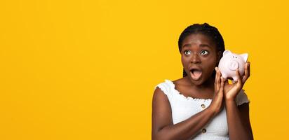 Shocked african american woman listening sound of coins in piggy bank, checking saved money, planning budget for future, emotional black lady standing over yellow studio background with copy space photo