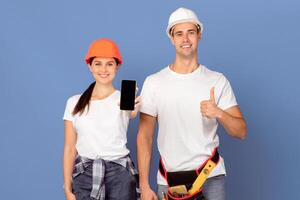 Cheerful couple of handyworkers in hardhats showing smartphone with black screen and gesturing thumb up, demonstrating copy space for your app or website design, standing over blue background, mockup photo