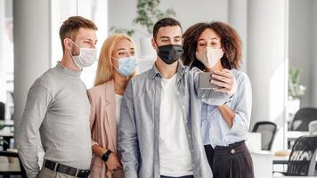Selfie or call for work. Colleagues of different nationalities stands together in protective masks and look at smartphone in guys hand, talking to boss on webcam, free space photo