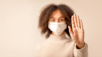 Stop worldwide COVID-19 epidemic and social distance. Focus on hand of african american woman in protective mask and blurred face of lady, isolated on light cream background, free space, studio shot photo