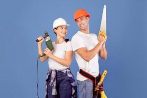 Ready For Repair. Smiling Couple In Protective Helmets Posing With Building Tools In Hands, Holding Saw And Drill, Standing Over Blue Studio Background, Enjoying Making Renovation Together, Copy Space photo