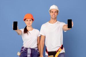 Smiling young man and woman in hardhats demonstrating smartphones with black screen, showing free copy space for your app or website design, standing over blue studio background, mockup image photo