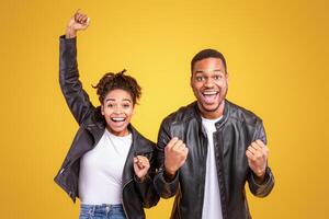 Joyful Black Spouses Gesturing Yes Shaking Fists In Excitement Posing Standing Over Yellow Background In Studio. Joy Of Victory, Winners And Happiness, Positive Emotions Concept photo