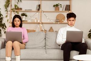 Distant Relationship. Unhappy Asian Couple Using Laptop Computers Sitting On Different Sides Of Couch Indoors, Separated By Distance. Long-Distance Relationship Problems And Difficulties Concept photo