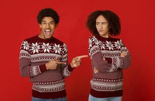 Relationship in couple and joke on holiday. Cheerful african americans guy with open mouth points his fingers at offended woman, crossed her arms on chest, isolated on red background, studio shot photo