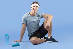 Full length portrait of young athletic man with dumbbells and bottle of water sitting on lilac studio background. Fit millennial sportsman looking at camera and smiling, resting after workout photo