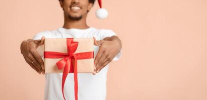Congratulations to New Year and Xmas. Smiling young african american man in white t-shirt giving gift box with present with red ribbon, isolated on light background, empty space, studio shot, panorama photo