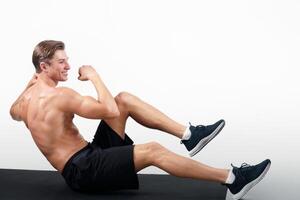 Happy young guy with muscular body working out on sports mat over light studio background. Strong millennial man with naked torso doing abs exercises, working out core muscles photo