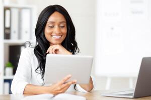 Joyful black business lady using tablet pc at workplace, speaking to colleagues on webcam, copy space. Successful millennial CEO reading important document online, checking email photo