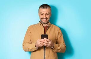 Cheerful Man Using Mobile Application On Cellphone Or Texting Via Smartphone Communicating Online Standing In Studio Over Blue Background. Communication And Technology photo