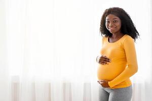 Beautiful pregnant african american woman hugging her big belly, enjoying her pregnancy, free space. Side view of young black expecting lady standing next to window at home, touching her tummy photo