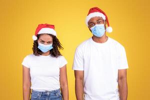 Coronavirus Christmas. Frustrated African American Couple Wearing Santa Hats And Protective Medical Masks Looking At Camera Standing On Yellow Studio Background. Xmas Party Cancelled Due To Covid-19 photo