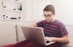 Teenager with computer on sofa at home. Boy engrossed in internet, typing on keyboard photo