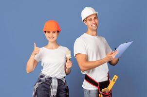 Repair Services Team. Happy Man And Woman In Hardhats With Clipboard And Paint Brush Posing Over Blue Background, Smiling Lady Showing Thumb Up, Ready For House Renovation, Copy Space photo
