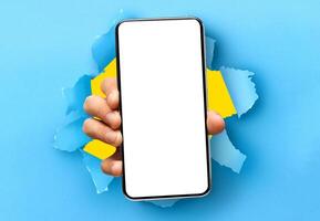 Mobile App Promo, Great Offer. Closeup of hand holding smartphone with white empty screen showing device close to camera breaking through blue paper sheet. Blank display with free copy space, mock up photo