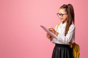 Apps for education. Girl pupil of elementary school in glasses in uniform and with backpack, typing on digital tablet, isolated on pink background photo