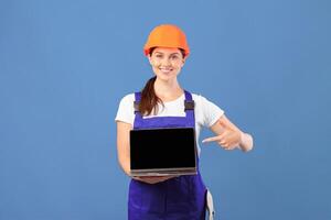 Smiling Lady In Coverall And Hardhat Pointing At Laptop With Black Screen, Recommending New Website For Online Shopping, Buying Construction Tools In Internet While Standing On Blue Background, Mockup photo